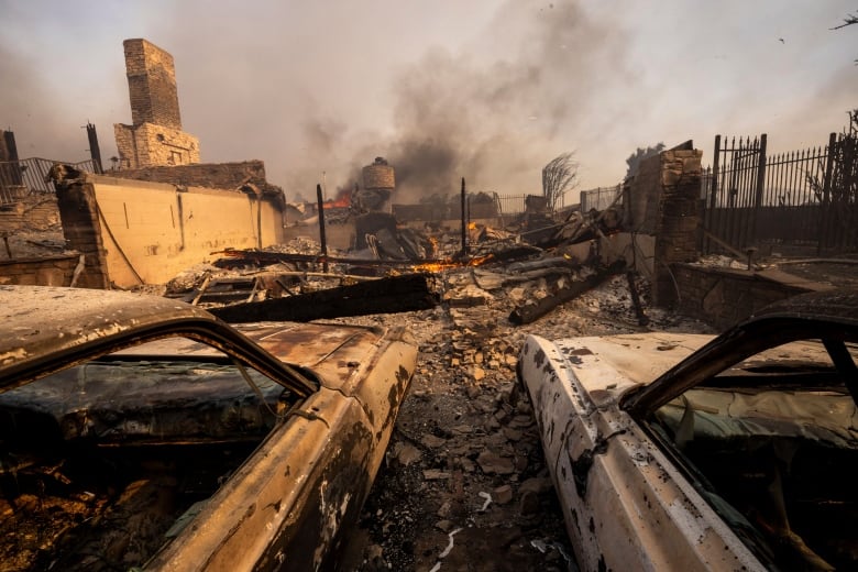 Charred vehicles are shown in the foreground on a debris-filled ground, as a burnt structure and smoke in the sky are shown.