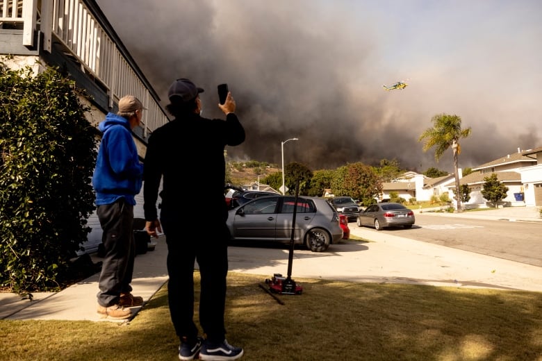 Two people wearing baseball caps with their backs to the camera stand on a suburban street lawn and hold their phones up, looking toward massive black smoke in the sky, with a helicopter in the skies.