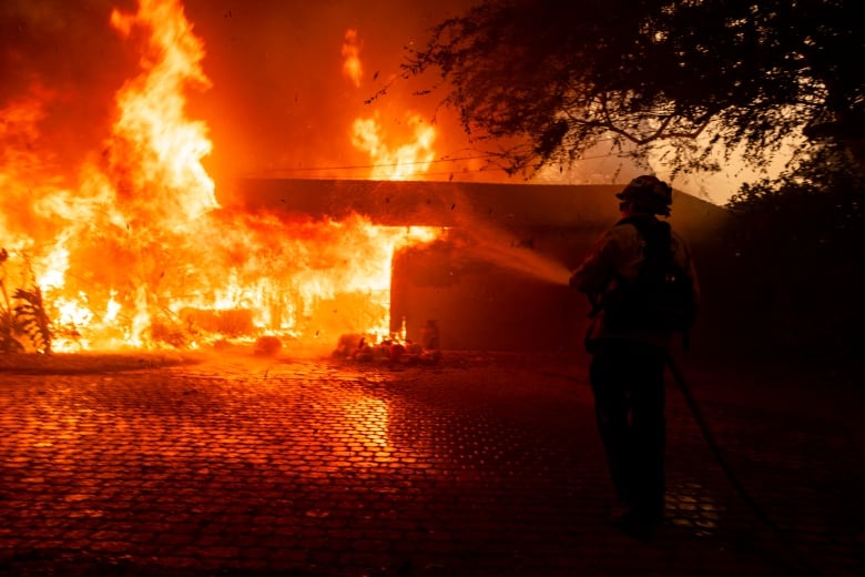 A firefighter is in the foreground with his back to the camera, spraying a hose toward a structure with massive orange flames surrounding it.