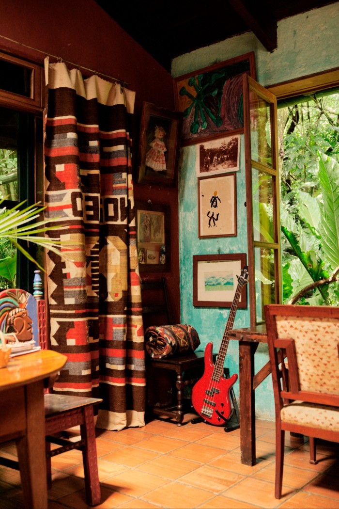 A room with a rustic turquoise coloured wall, small table and chairs, terracotta tiled floor and dark wooden beams overhead. An electric bass guitar leans against the wall. Above it are framed artwork and paintings. On the left, there’s a thick, patterned curtain with tribal-inspired designs