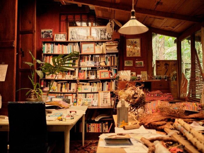 A cozy workspace filled with books, art supplies, and natural elements. Windows to the right provide a view of the greenery outside