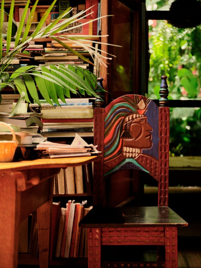 A wooden chair with intricate carvings and a colourful painted design of an indigenous figure is positioned in front of a bookshelf overflowing with books 