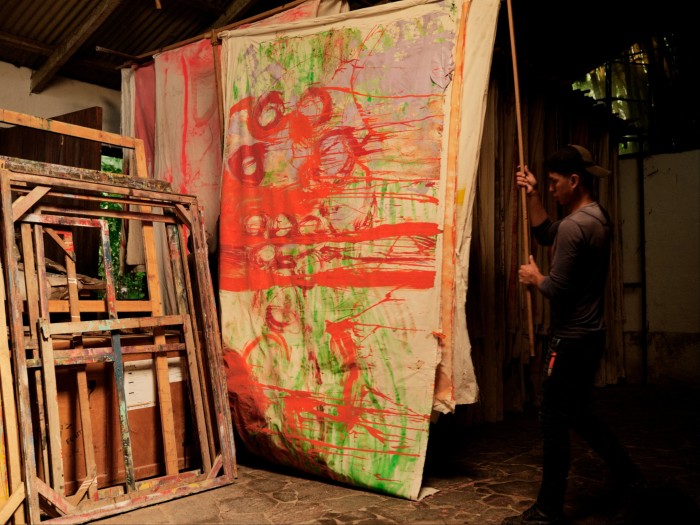 An artist’s studio with a large, abstract painting on a canvas that is being moved or adjusted by a person. The canvas is painted with bold, expressive strokes in vivid colours