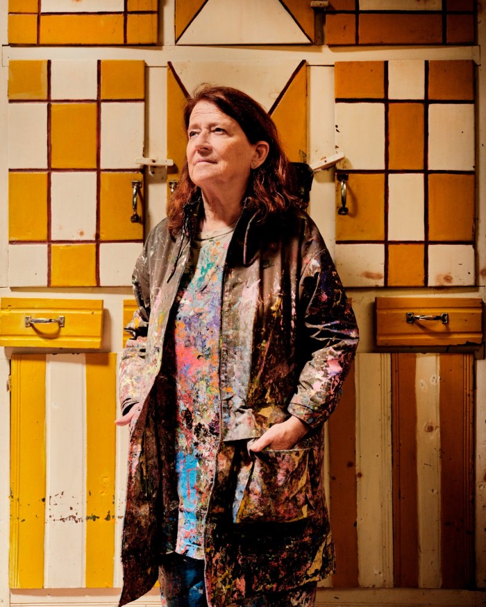 A woman dressed in a long, paint-splattered jacket is standing against a background of wooden cabinets painted with a geometric pattern in shades of yellow, white, and brown