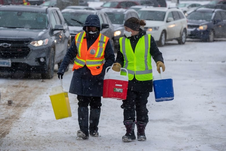 Two people in winter clothing, including coats, mittens and medical masks carry coolers past vehicles on a grey, snowy day.