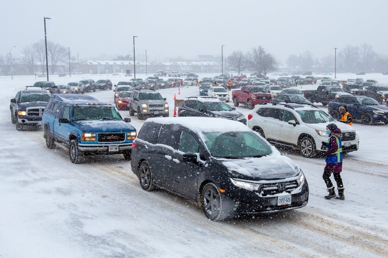 Long lines of vehicles are shown arriving at a drive-thru vaccination clinic on a grey, snowy day.