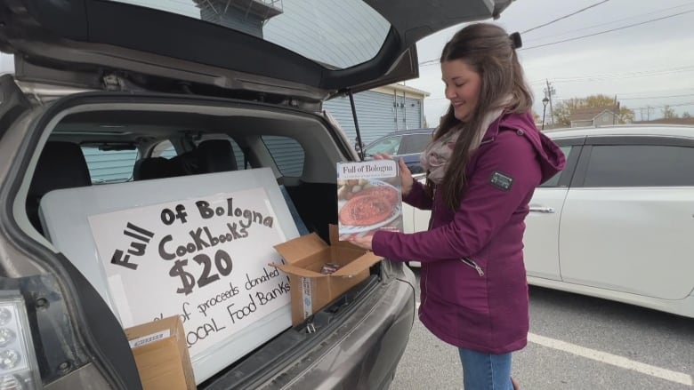 A woman stands at the back of her car in a parking lot, smiling and holding up a book called Full of Bologna. In her trunk are boxes of books and a big sign that reads: Full of Bologna Cookbook $20"