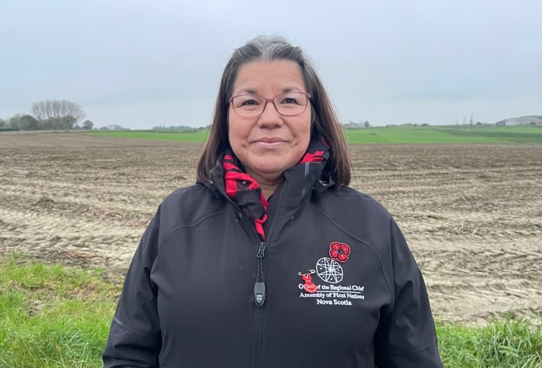 A woman stands in a field, looking at the camera, smiling slightly. 