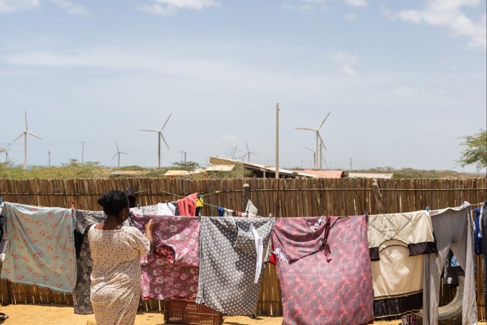 A woman hanging laundry on a clothesline in a rural setting. In the background, multiple wind turbines rise above low, sparse vegetation and small structures