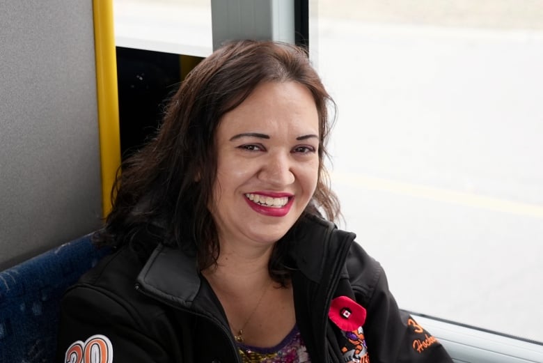 A woman with shoulder length dark hair, red lipstick and a dark jacket sits in a bus set by the window smiling at the camera.  