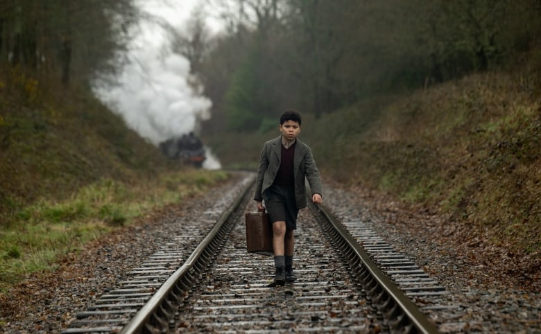 A young boy walks along train tracks. Behind him is an oncoming train. 