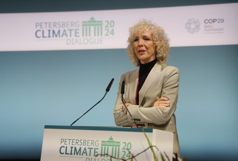 A woman in a beige suit speaks at a podium.