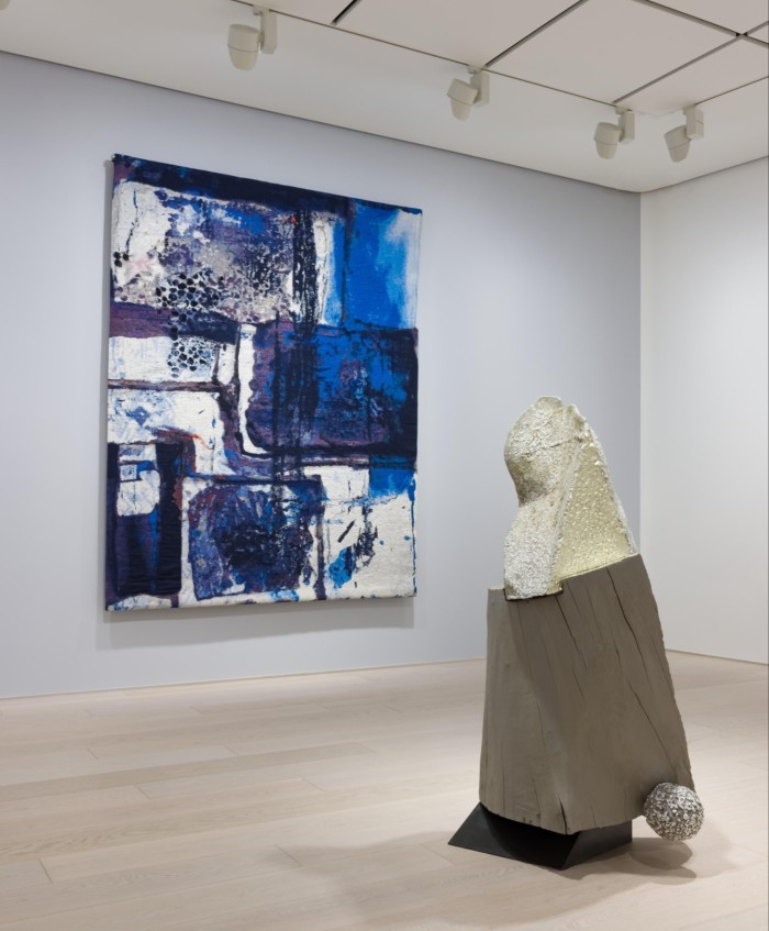 An art gallery has on its wall an abstract painting in blocks and blotches of blue paint; on the floor is a sculpture in mixed materials