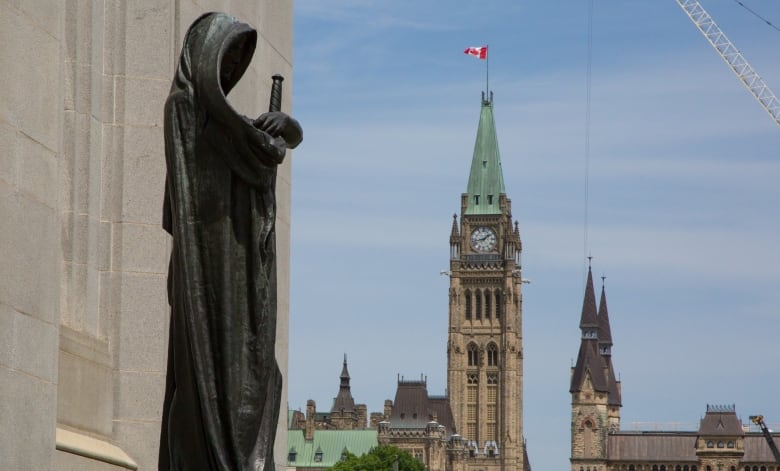 A statue of a cloaked figure, holding a sword, stands at the front of a building. The spires of other buildings are visible in the background. 