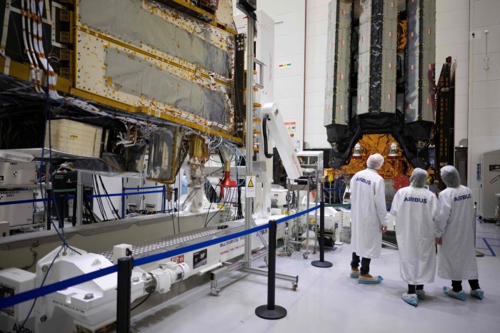 Airbus employees, wearing white lab coats with the company's logo, stand in a clean room observing the meteorological satellites.