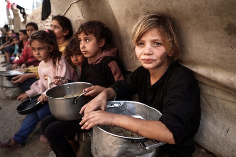 Children sit on a bench in a tent holding tin pots.