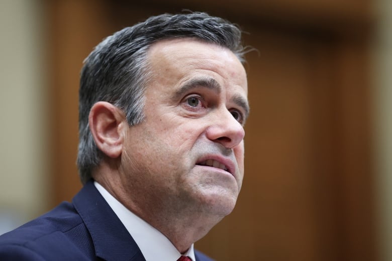 A cleanshaven man with dark hair with streaks of grey, wearing a suit and tie, speaks in a closeup photo that is taken indoors.