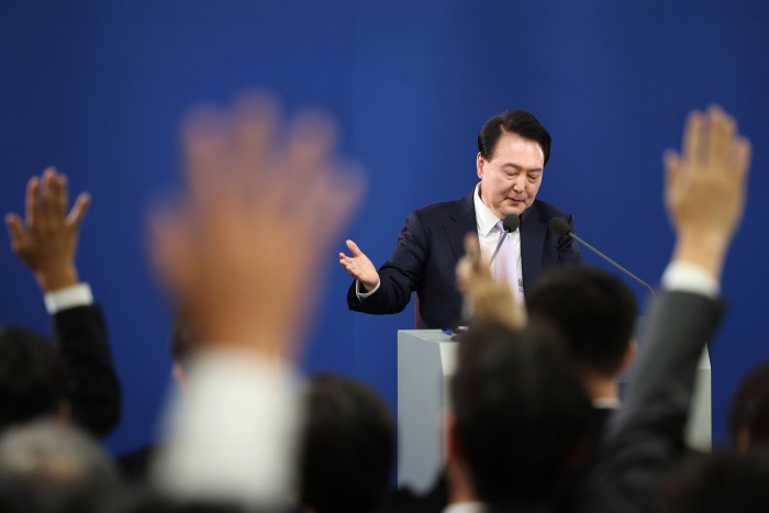 A man in a suit, standing at a podium with two microphones, appearing to address an audience