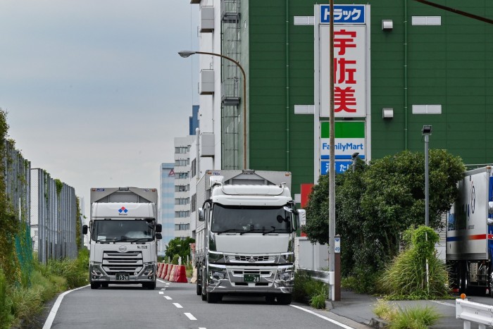 Two large trucks driving on a narrow road lined with greenery, with a green building and a sign featuring Japanese text and a FamilyMart logo in the background