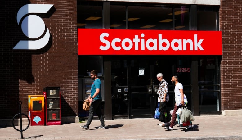 People walk past a brick building that says Scotiabank in white letters on a red banner. The people are in t-shirts. 