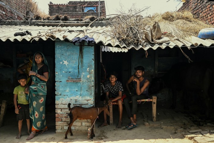 A family outside their rustic home, with a woman in a green sari standing beside a young boy, while two young men sit on a traditional bench. A goat stands near them, and the roof is covered with dried vegetation