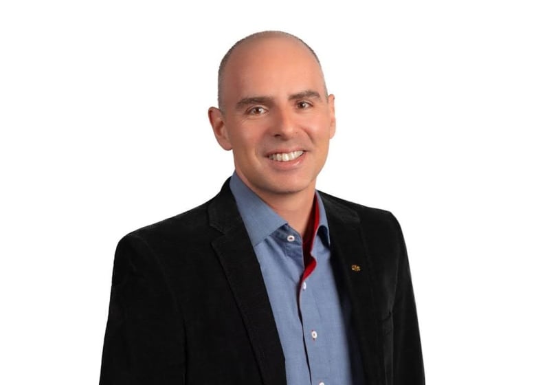 A white man with a shaved head, a blazer and blue button-up stands in front of a white background.