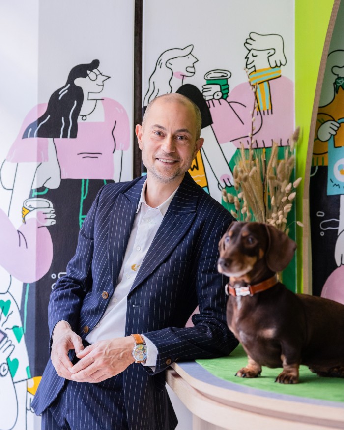 A man in a pinstripe suit leans casually against a counter. Beside him on the counter sits a small brown dachshund with a leather collar