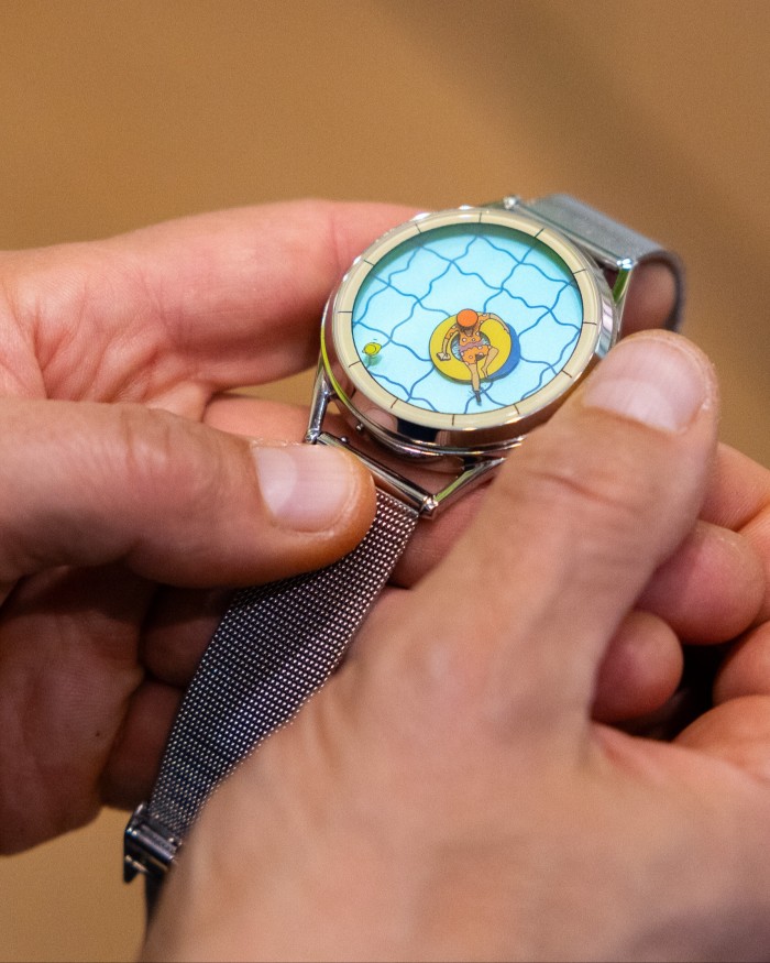 A pair of hands holding a watch that features a man floating on an inflatable ring in a swimming pool, with one foot acting as the hour hand and a rubber duck reading off the minutes