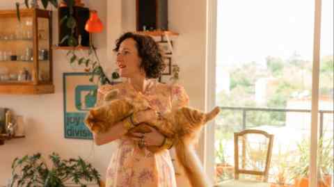 A woman in a long floral dress stands in a light-filled room holding her large ginger cat