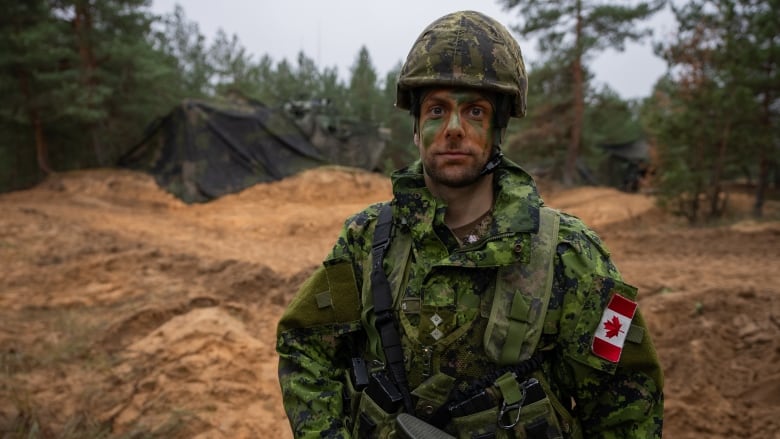 A soldier in green camouflage fatigues and face paint stands in a field. 