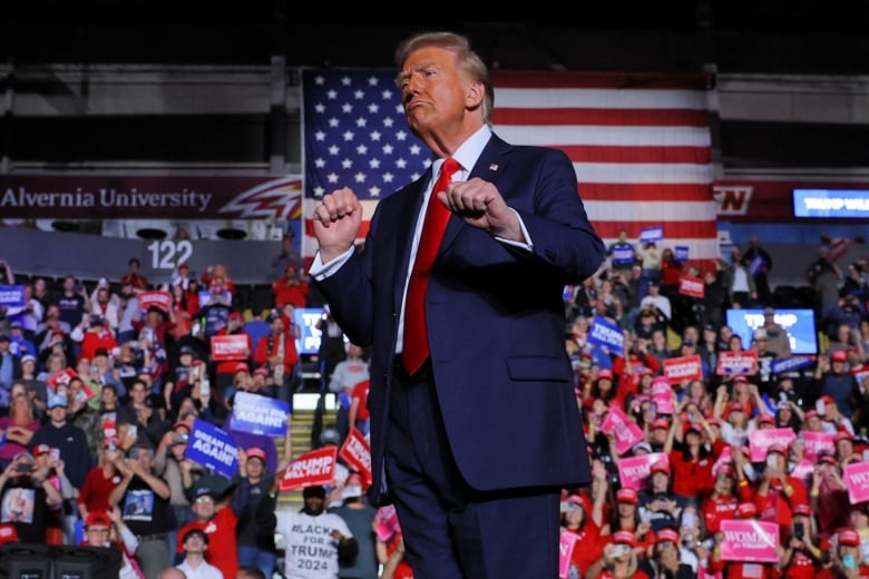 A man in a blue suit and red tie gestures in front of a crowd of people.