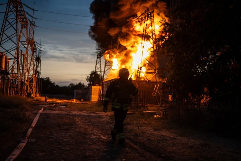 A large orange fire is shown near a hydro tower as a firefighter in uniform is shown in the foreground in shadow.