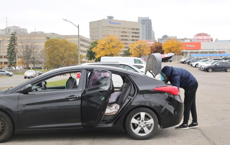 A man wearing a dark blue ski jacket and black pants rummages in the trunk of his dark-coloured car.