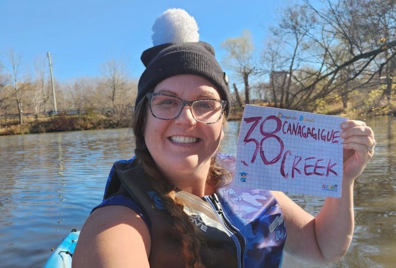 Woman in kayak takes selfie while on a lake. The sign she's holding says "38" and Canagagigue Creek.