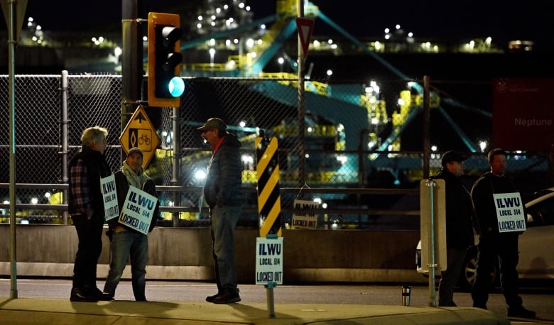 A nighttime scene shows five workers with signs around their neck that say "ILWU Local 514 Locked Out" standing on the pavement. A big teal crane structure is visible behind the fence behind them. 