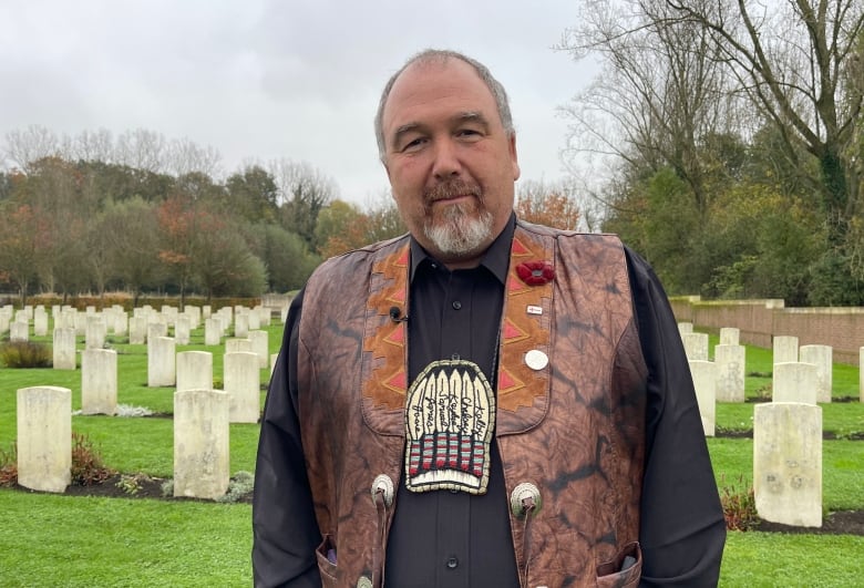 A bearded man, wearing a leather vest, stands in a cemetary. 