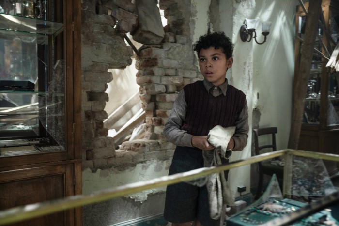 A young mixed-race boy stands close to a bombed-out house