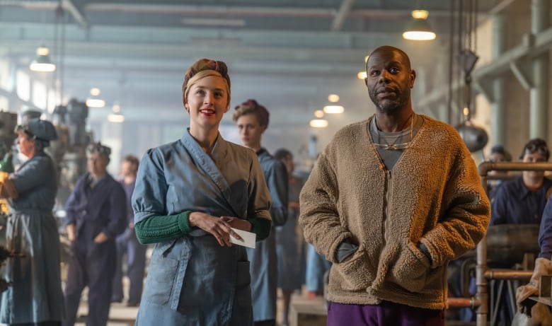 A woman wearing a blue factory outfit stands next to a man wearing more modern clothes. Behind them are other factory workers.