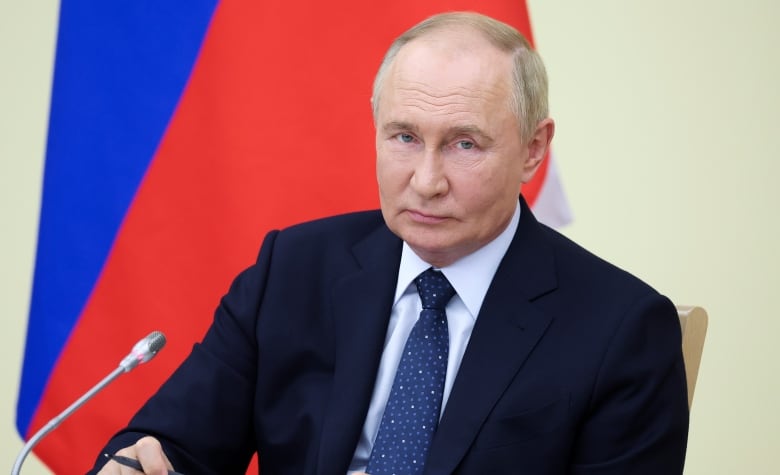 A man in a suit, seated at a meeting table, looks at the camera. A blue and red flag stands behind him. 