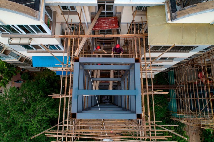 Workers wearing red helmets install a lift shaft in an old residential building. The structure is surrounded by scaffolding, and the view is from above, showing the construction in progress