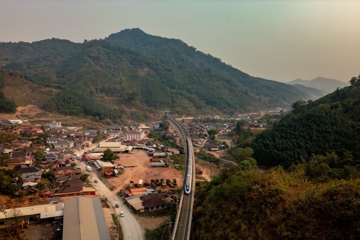 An aerial view of a rural town surrounded by green hills and mountains, with a modern train line cutting through the center of the community