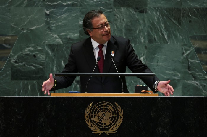 A man wearing a dark suit with a white shirt and a red tie is speaking at a podium with the emblem of the United Nations in front of him, indicating that he is addressing an audience