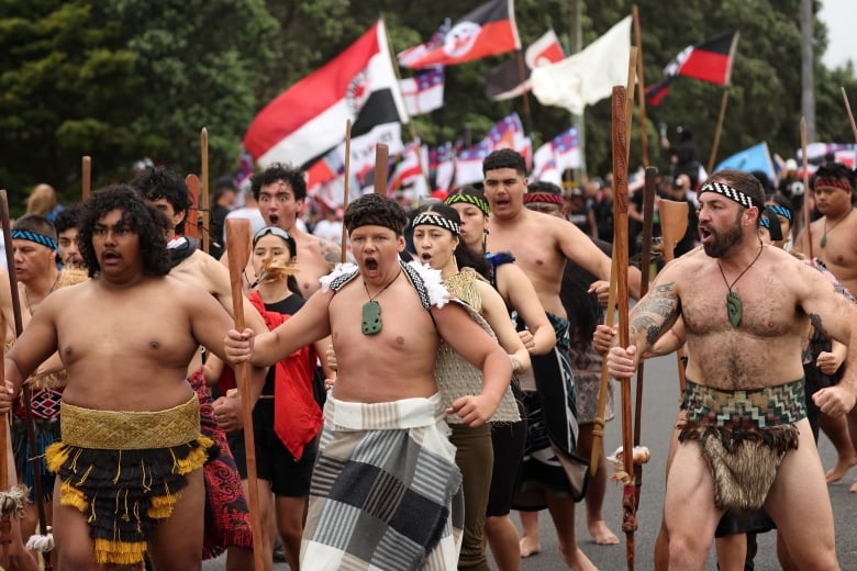 Several shirtless men in loin cloths holding what appear to be spears are shown chanting and walking in an outdoor protest photo.