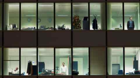 A view from outside of two office floors at night. We can see a Christmas tree, one man sitting at a desk and two men gazing out of the windows