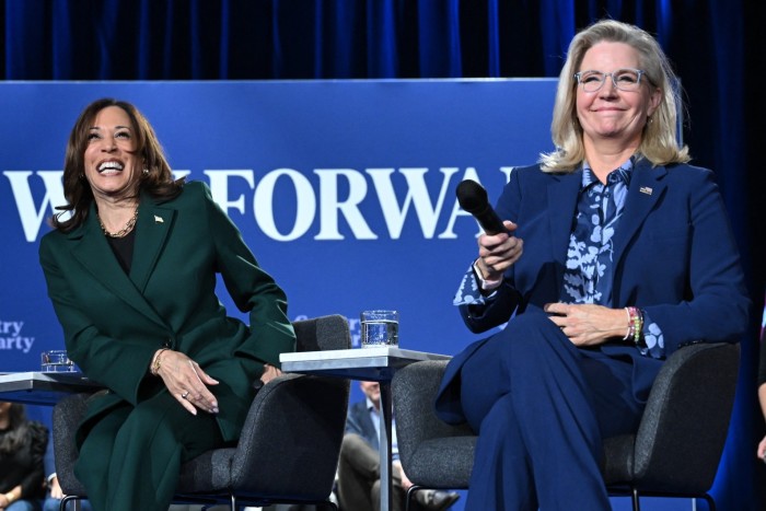 Kamala Harris and Liz Cheney sit on stage during a town hall discussion. Harris is wearing a green suit and laughing, while Cheney, in a blue suit, holds a microphone and smiles.