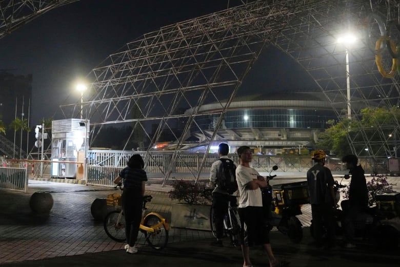 Several people are shown on the sidewalk outside an arena-like building in a nighttime photo.