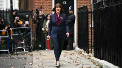 Rachel Reeves walks outside 11 Downing Street in London holding the red Budget Box. Photographers and a police officer are visible in the background