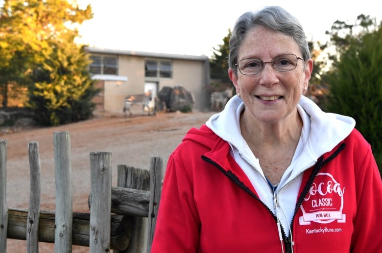 A woman with short, grey hair and glasses wearing a red jacket and white blouse stands in front a wooden fence.