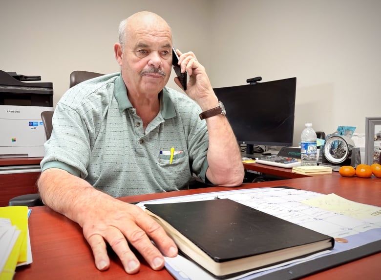 A man in a celadon polo shirt  speaks on the phone while sitting at a desk.