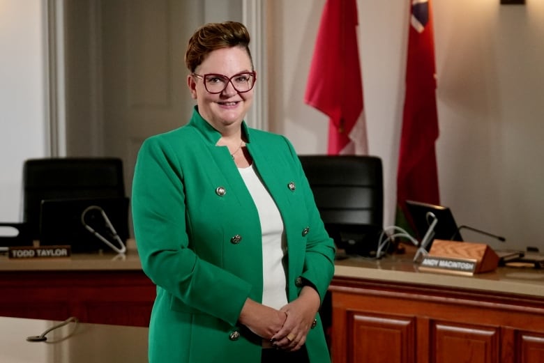 A woman with short red hair and glasses wearing a green jacket with silver buttons and a white shirt stands in front of the Ontario and Canadian flags.   